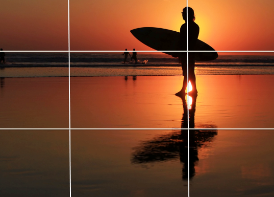 Une photographie d'un surfeur debout sur la plage, tenant sa planche, illustrant la composition d'une image selon la règle des tiers, par SmartCuts Creative, basé à Bussigny et Lausanne, en Suisse.