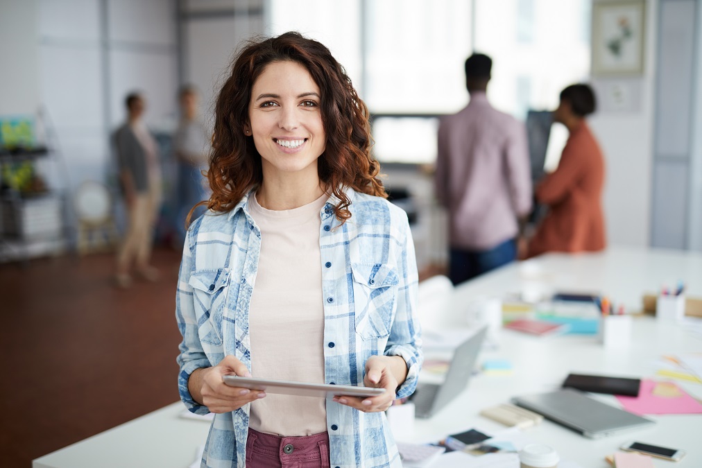 Waist up portrait of young woman smiling and holding digital tablet while standing in the SmartCuts Creative in Geneva and Lausanne, Switzerland.